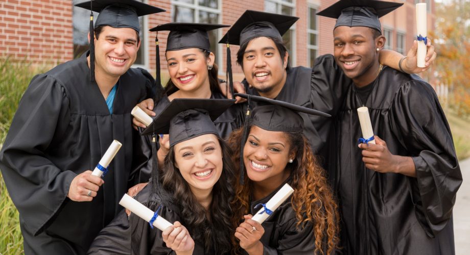 Six multi-ethnic friends dressed in cap and gowns excitedly show off diplomas after college graduation. School building background.